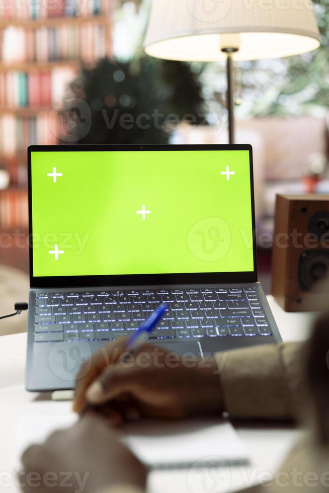 Entrepreneur using a notebook to write notes next to green screen display on laptop, working from home to help develop the startup. Businessman analyzes accounting data at desk. photo