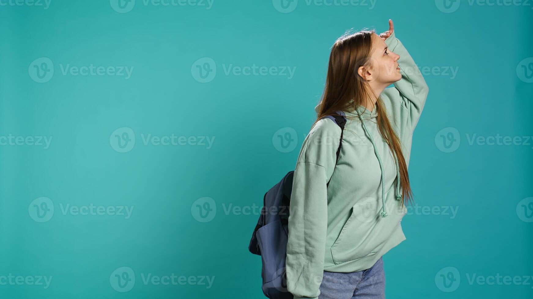 Curious smiling girl holding rucksack looking around for something, doing hand gesturing. Upbeat person doing surveillance gesture, exploring, seeking things, isolated over studio backdrop, camera B photo