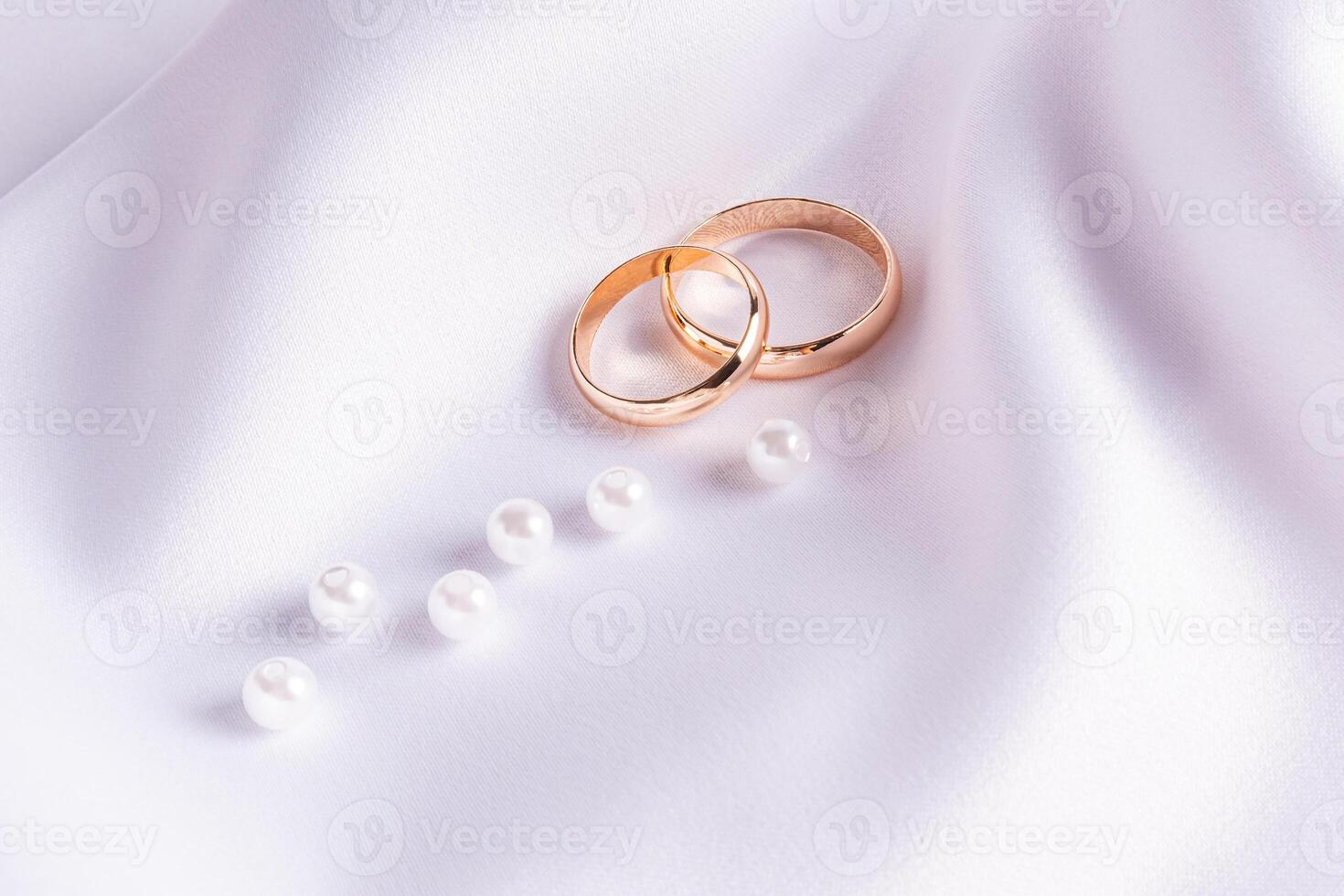 A pair of gold wedding rings on a white satin background with a scattering of several pearls. postcard. invitation. A copy space. minimalism. photo