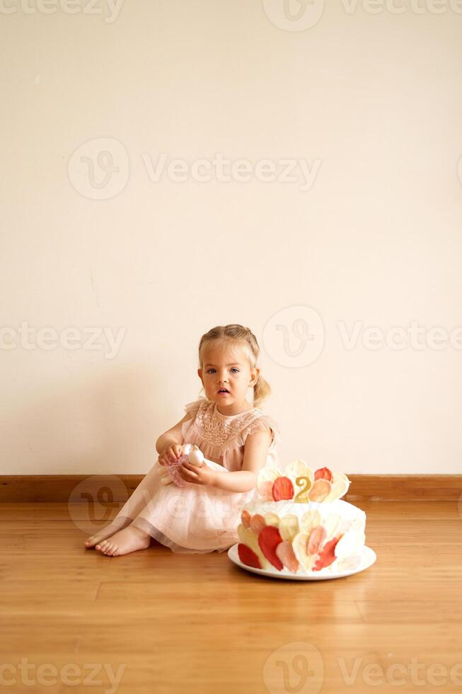 Little girl sits on the floor near a white chocolate cake with colorful decorations on a plate photo