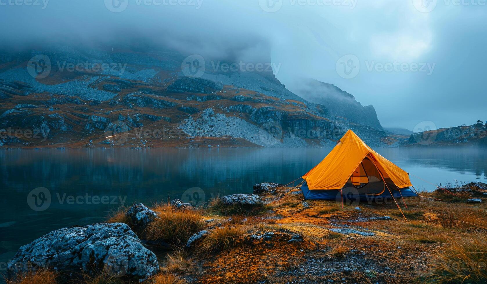 A yellow tent is set up on a rocky shore near a lake. The sky is cloudy and the water is calm photo