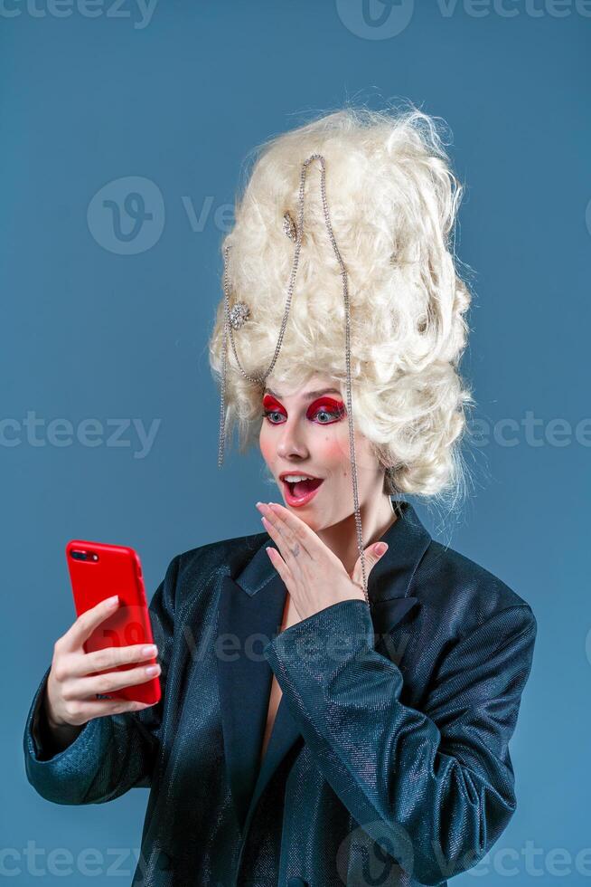 Amazed woman in makeup and royal wig using smartphone in blue studio photo