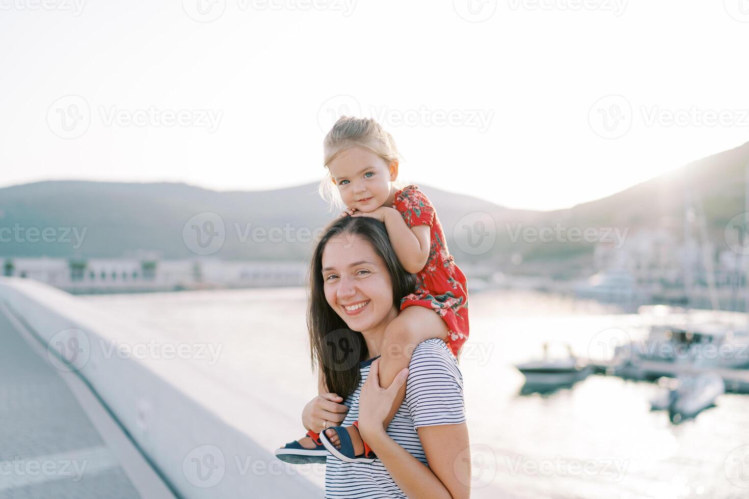 Little girl sits on the shoulders of a smiling mother, resting her chin on her head on the seashore photo