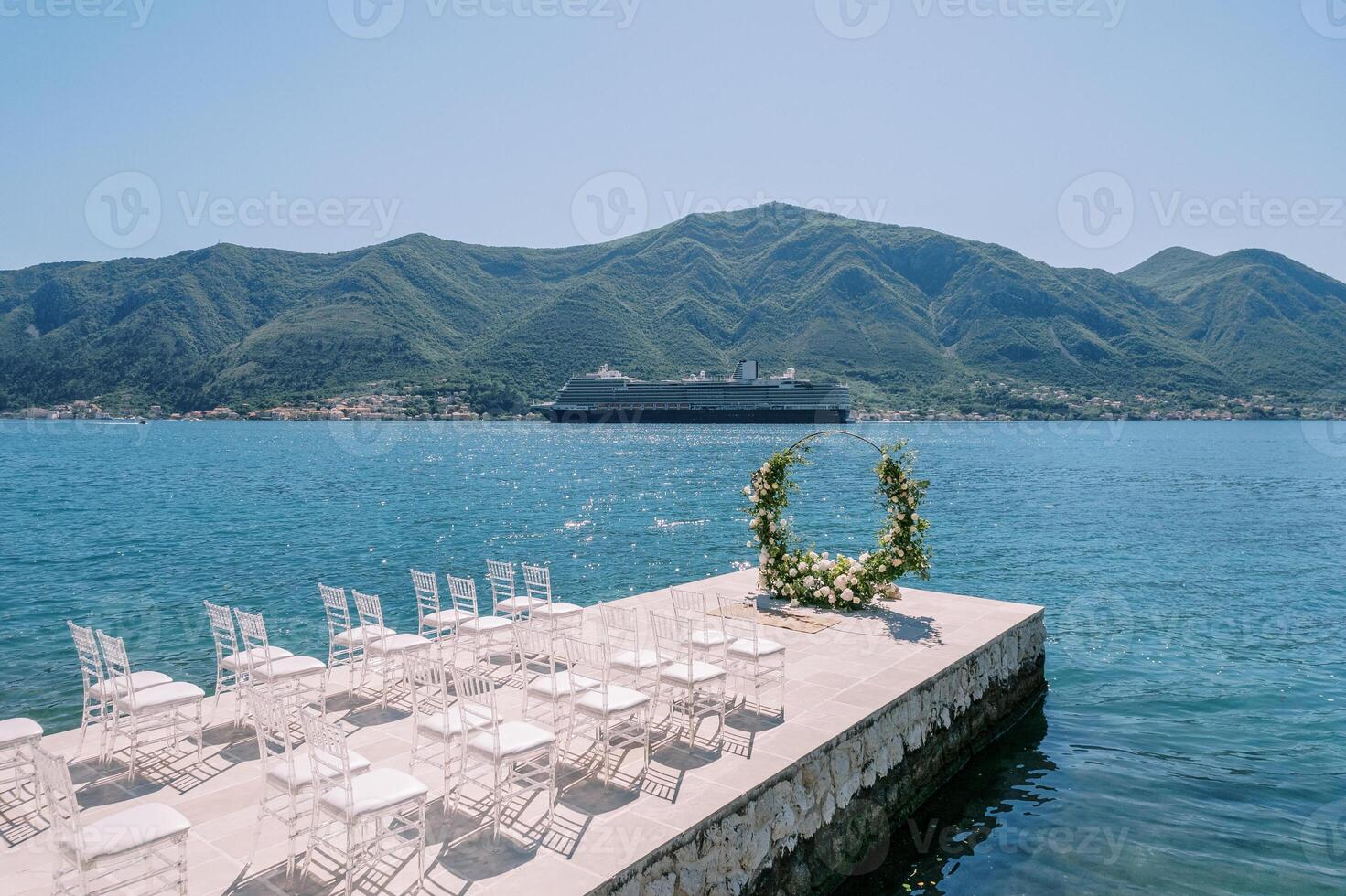 White chairs stand in front of a round wedding arch by the sea, with a cruise ship in the background photo