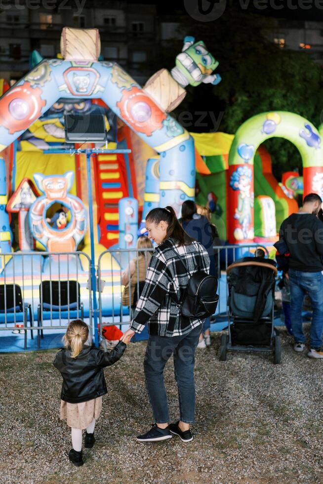 Mother and a little girl stand holding hands in front of a colorful attraction. Back view photo
