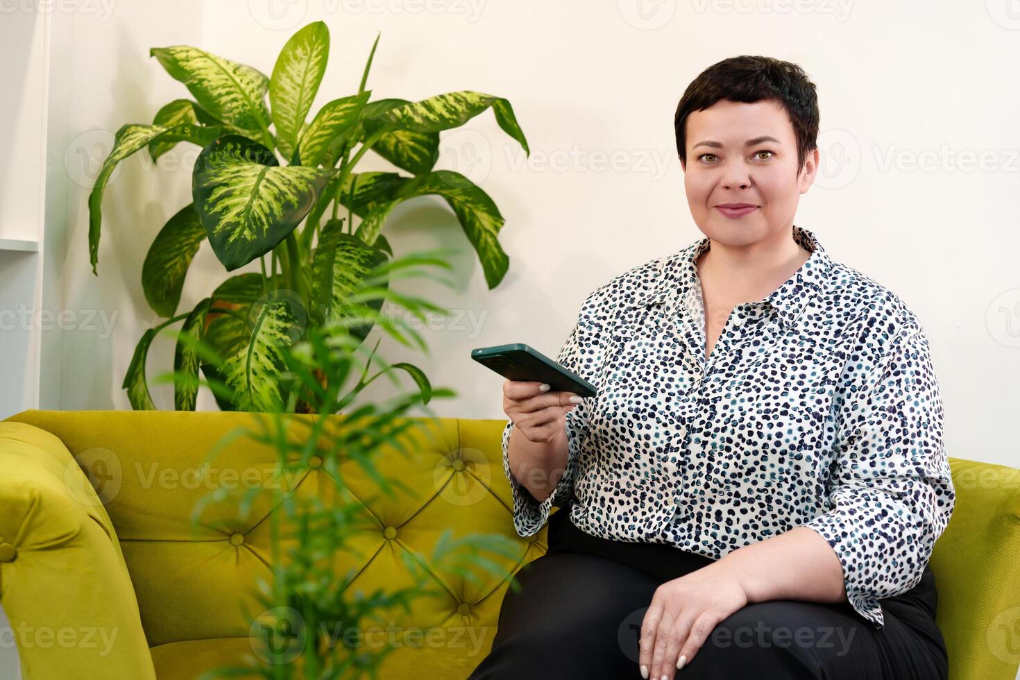 Successful and goal-oriented middle-aged woman demonstrating business proficiency while talking on the phone while sitting comfortably on a sofa in a modern office photo