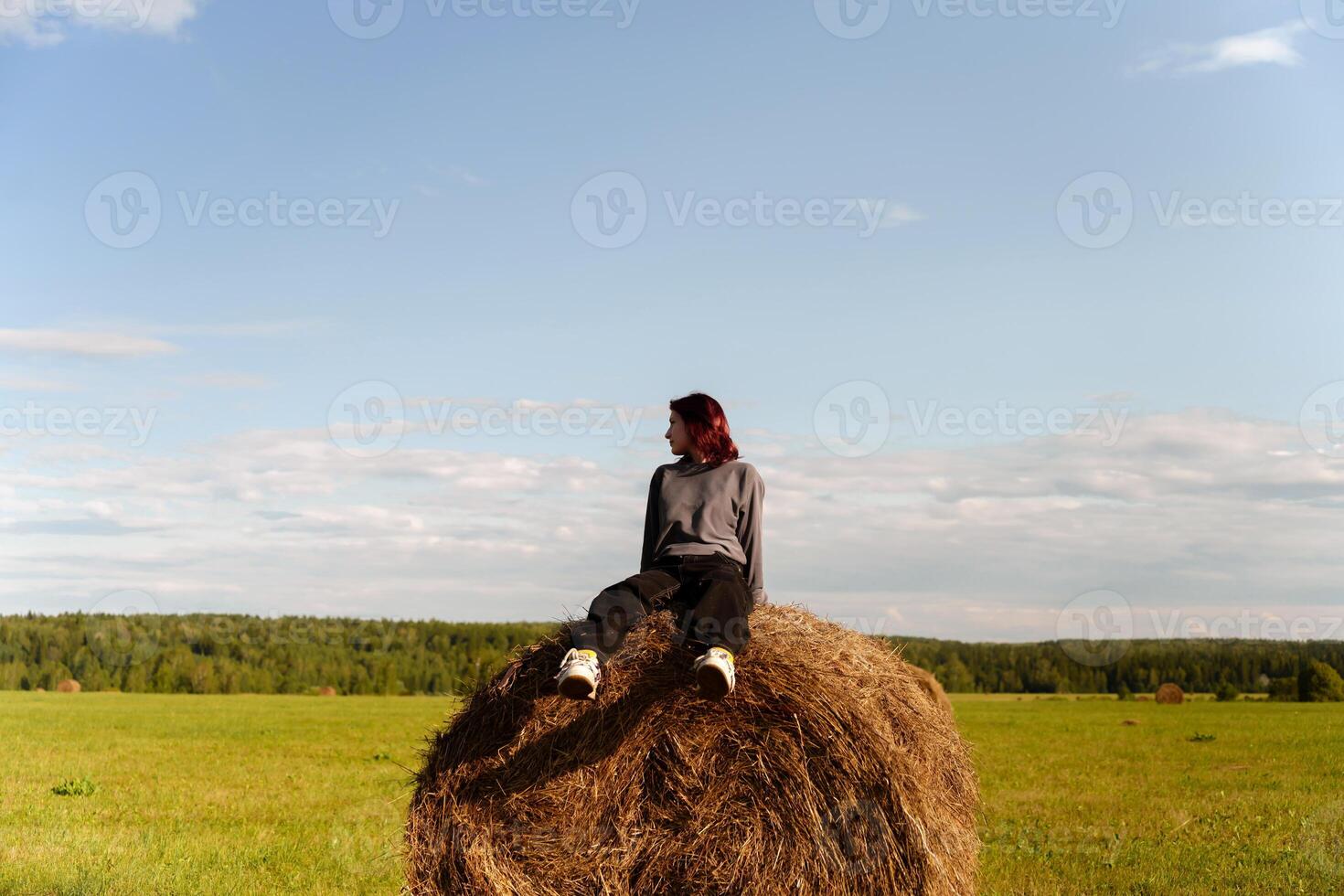 Rebellious teen in modern over-size clothing sits in a field on a haystack with a blue sky overhead photo