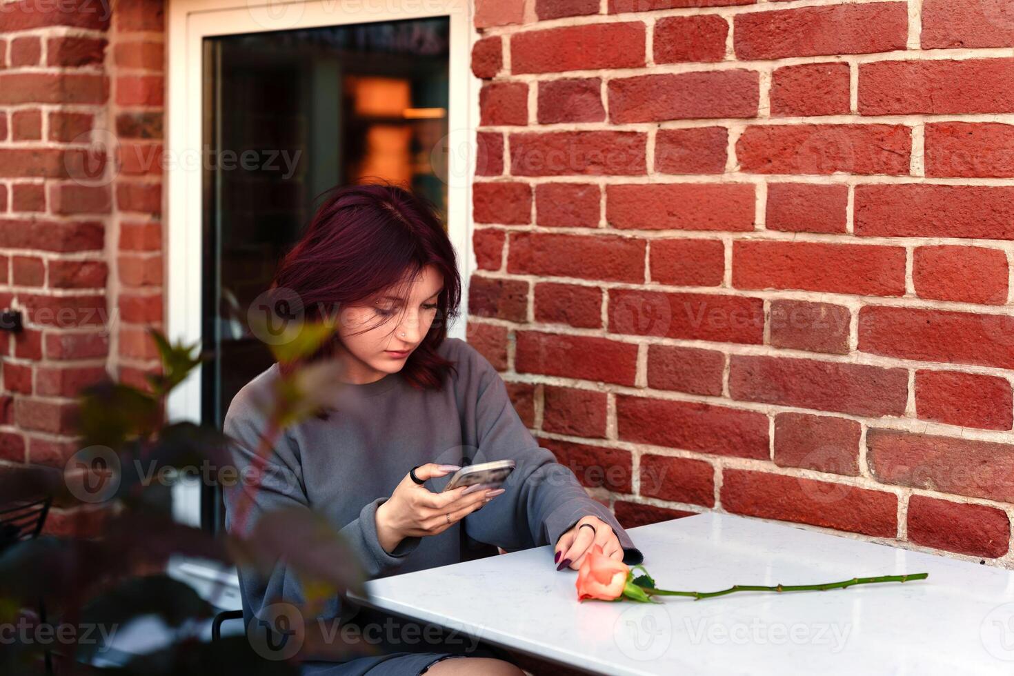 troubled teen with dyed hair using her cell phone screen sitting at a table in a cafe against a brick wall, a lone rose complements the pensive scene photo