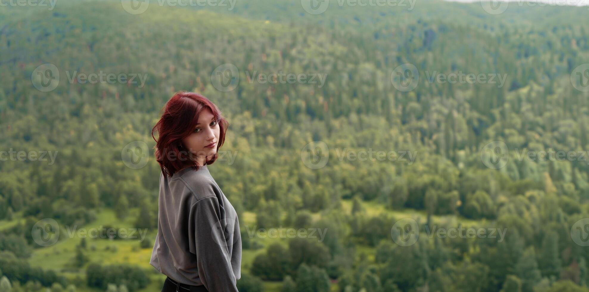 zoomer girl with dyed hair and facial piercings stands on top of a mountain against the backdrop of a dense green forest photo