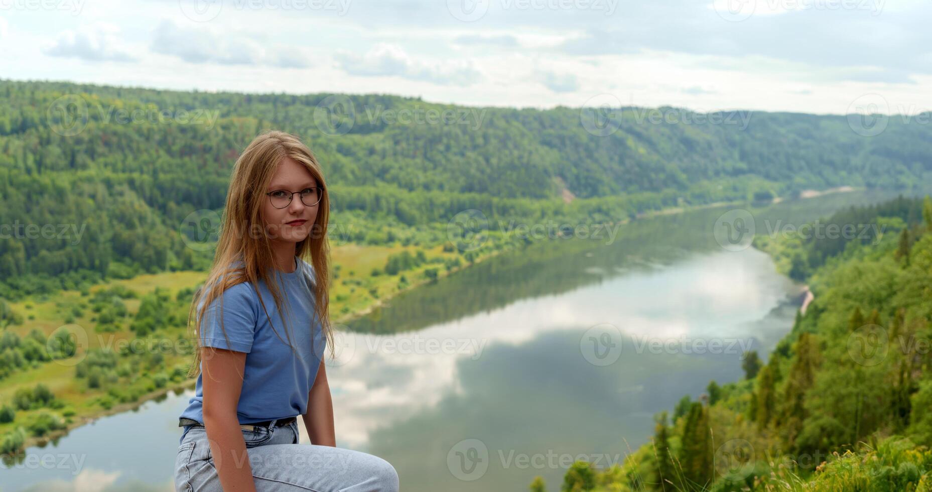 young, blonde-haired girl stands boldly against the backdrop of a majestic mountain, wearing a simple blue tee and blue jeans, with ample space for text overlay photo