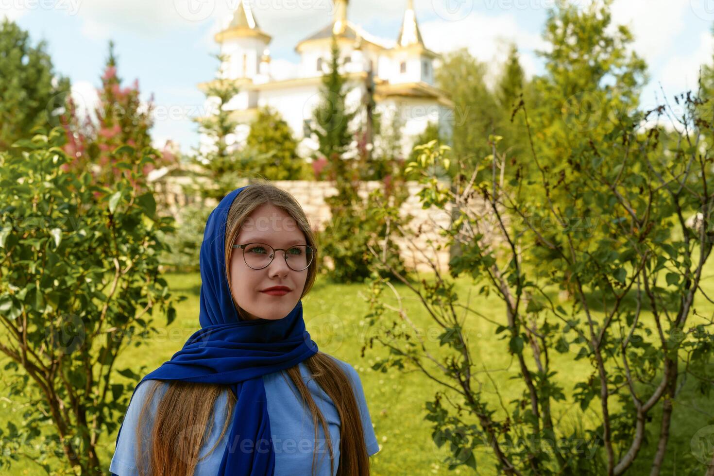Portrait of teenager in blue headscarf and gold cross on a chain, against the backdrop of a white church with golden domes, conveying Orthodox Christian essence photo