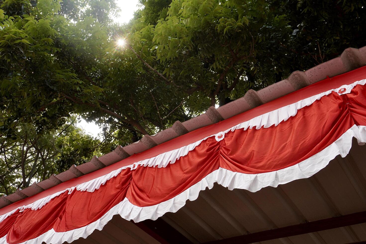Red and white flag decoration hanging on building roof isolated on horizontal outdoor green tree leaves and bright sky background. Bendera merah putih fabric cloth texture. photo