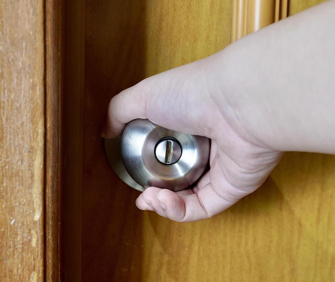 Human hand with light skin tone with opening door gesture isolated on horizontal brown door with round cylinder metal bathroom knob background. photo