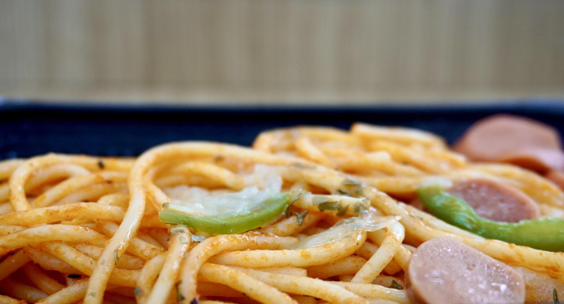 Close up cooked spaghetti noodles with tomato sauce and sausage slices isolated on horizontal black plate and wooden wall background. photo
