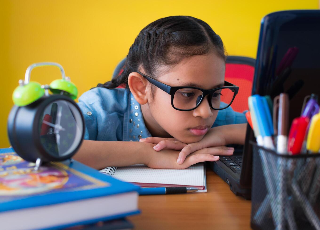 Cute girl wearing glasses is searching for information on internet from laptop on the desk isolated yellow background, Education concept. photo