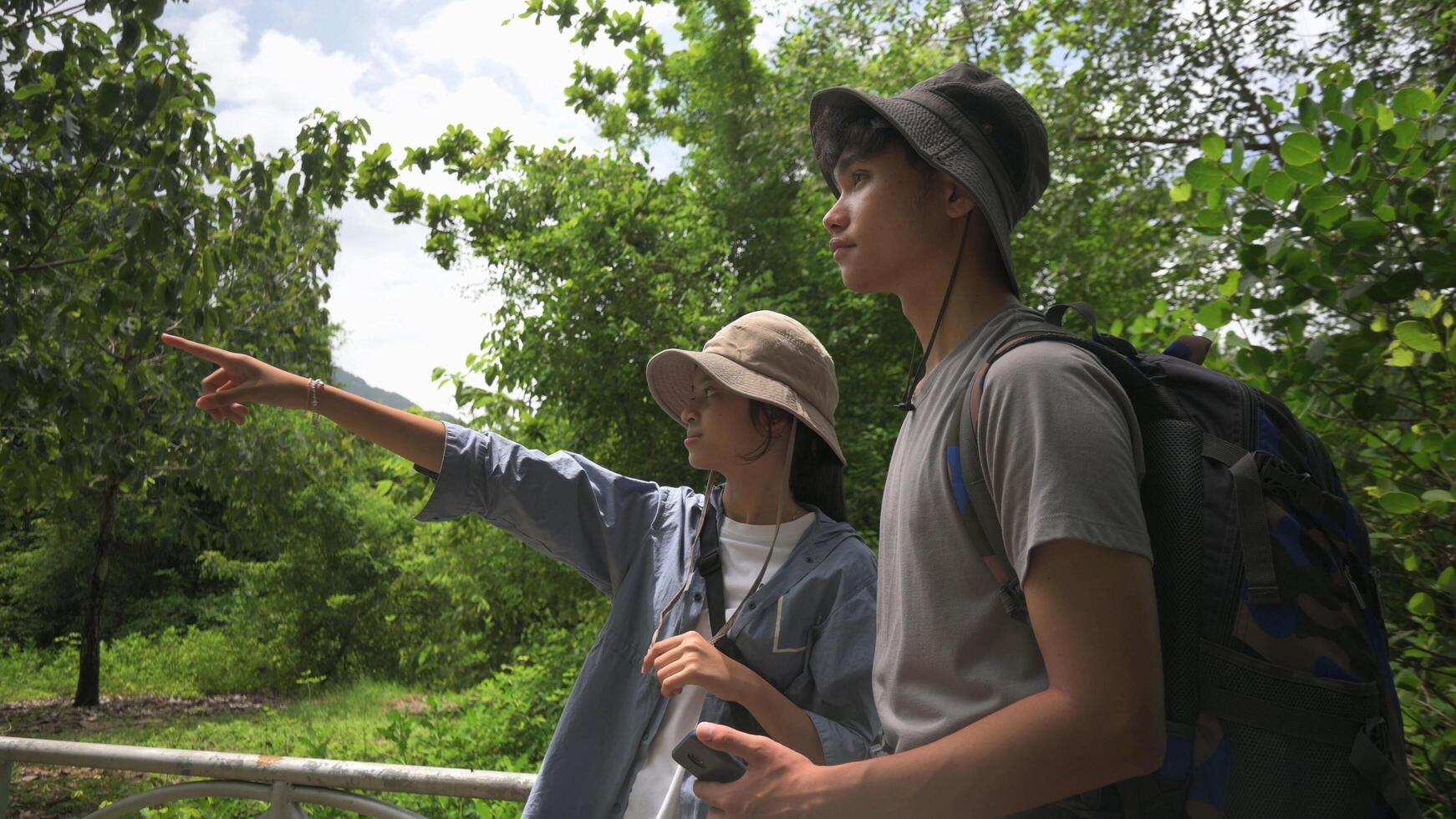Two Asian young teen backpackers looking for directions while hiking in tropical forest park in summer holiday. Outdoor pursuits. Teenager hobbies and leisure activities. photo