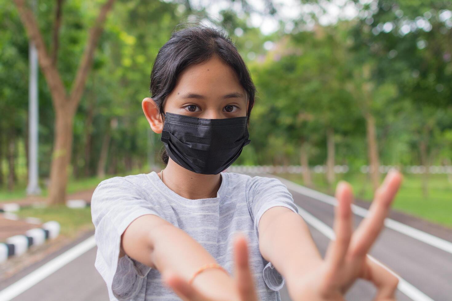 Portrait Asian pretty teen girl in casual dress wears black protective mask posing gesturing with her fingers while standing on the road in public park during COVID-19 epidemic. photo