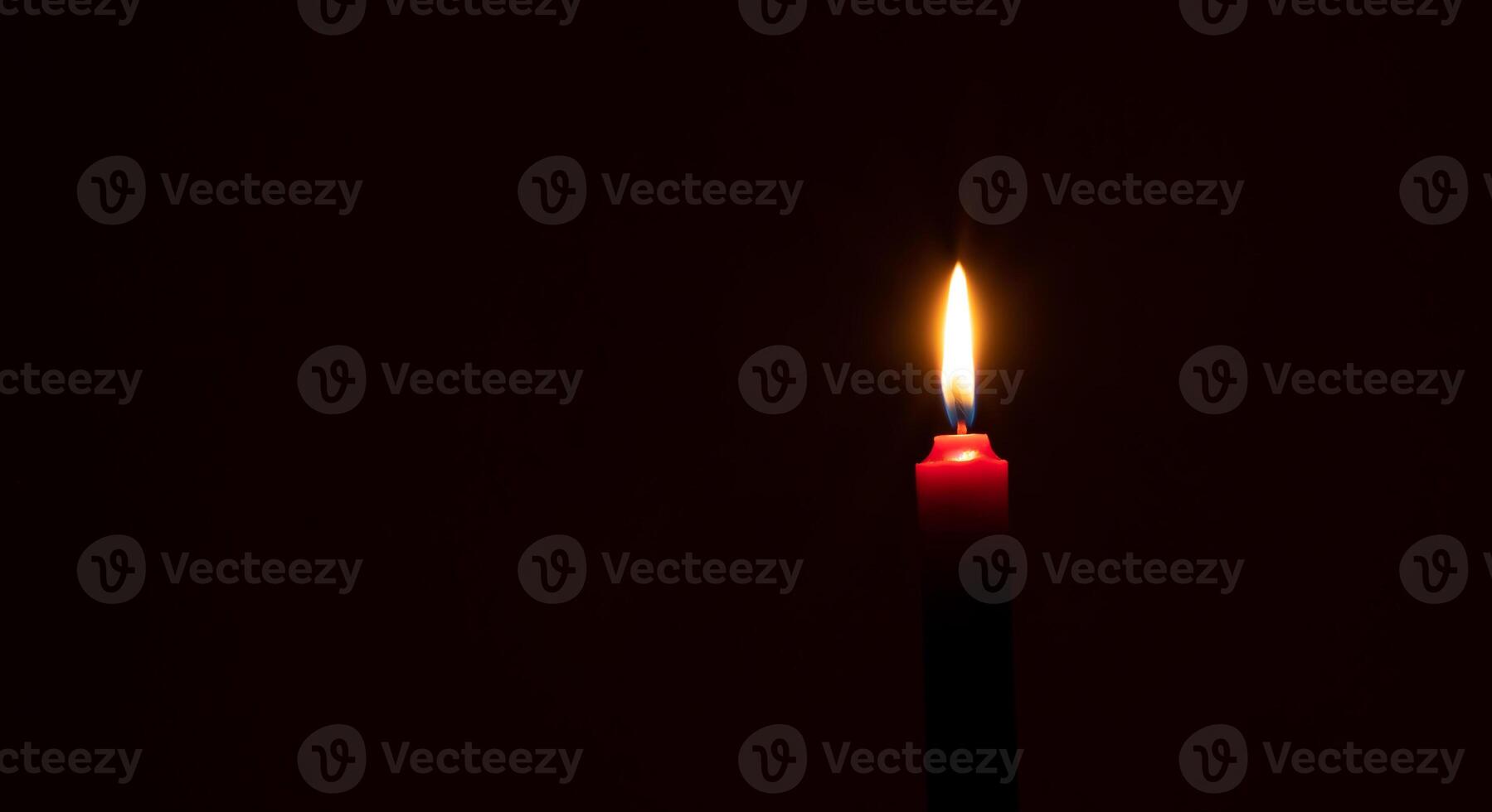 Single burning candle flame or light glowing on a small red candle on black or dark background on table in church for Christmas, funeral or memorial service with copy space. photo