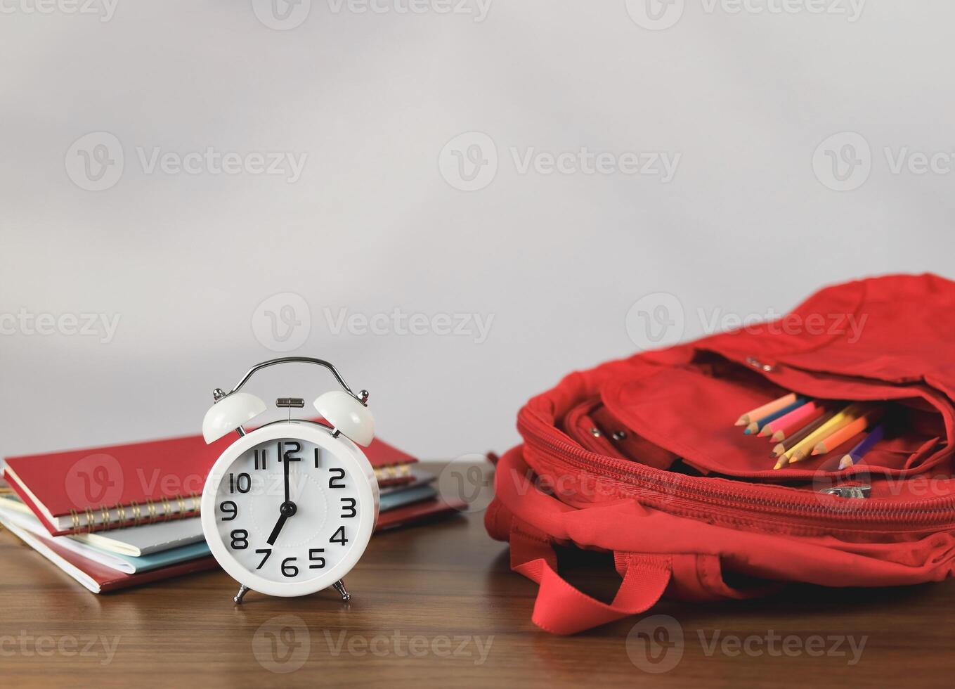 red school backpack with stationery, books and white alarm clock 7 o'clock on wooden table , white wall. photo