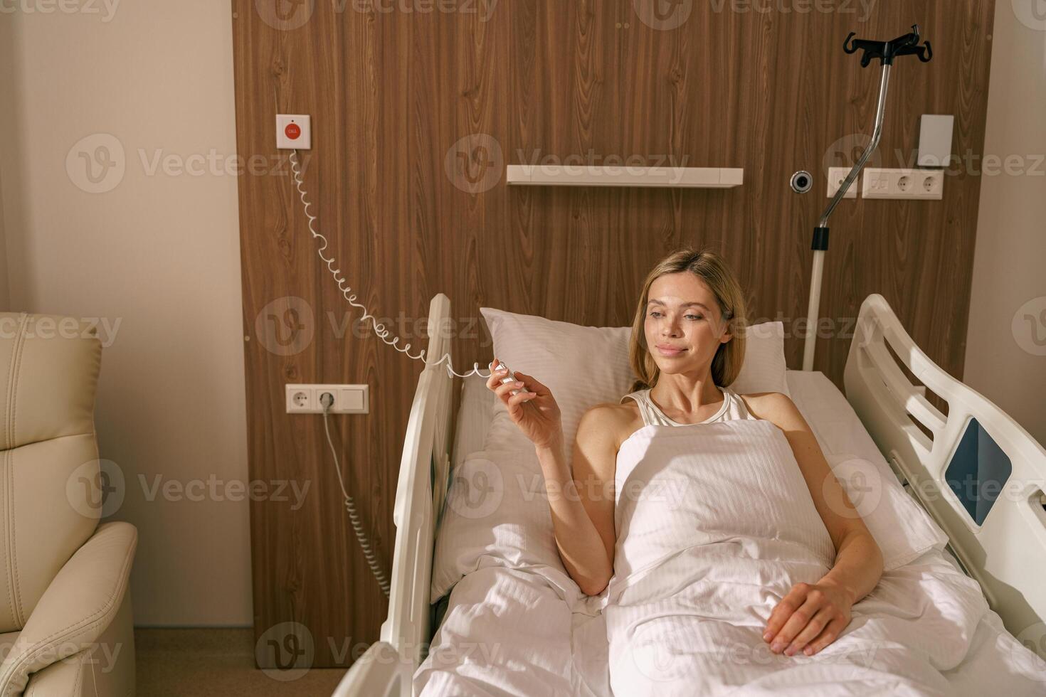 Young smiling woman patient holding emergency call button while lying in hospital bed photo