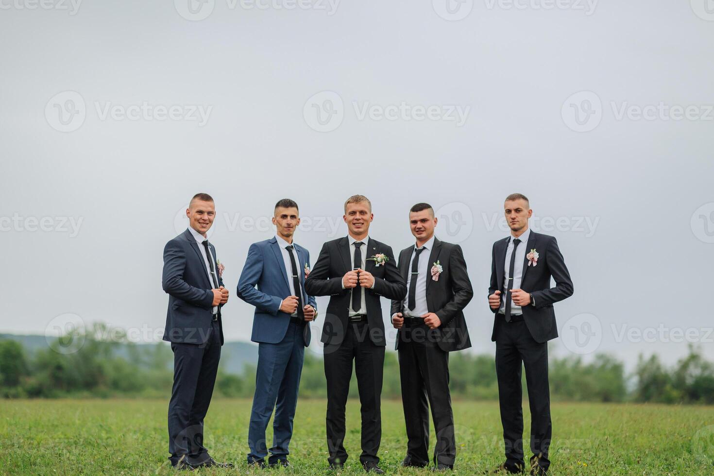 A group of five men are standing in a field, all wearing suits and ties. They are posing for a photo, with one man holding a wine glass. Scene is formal and elegant photo