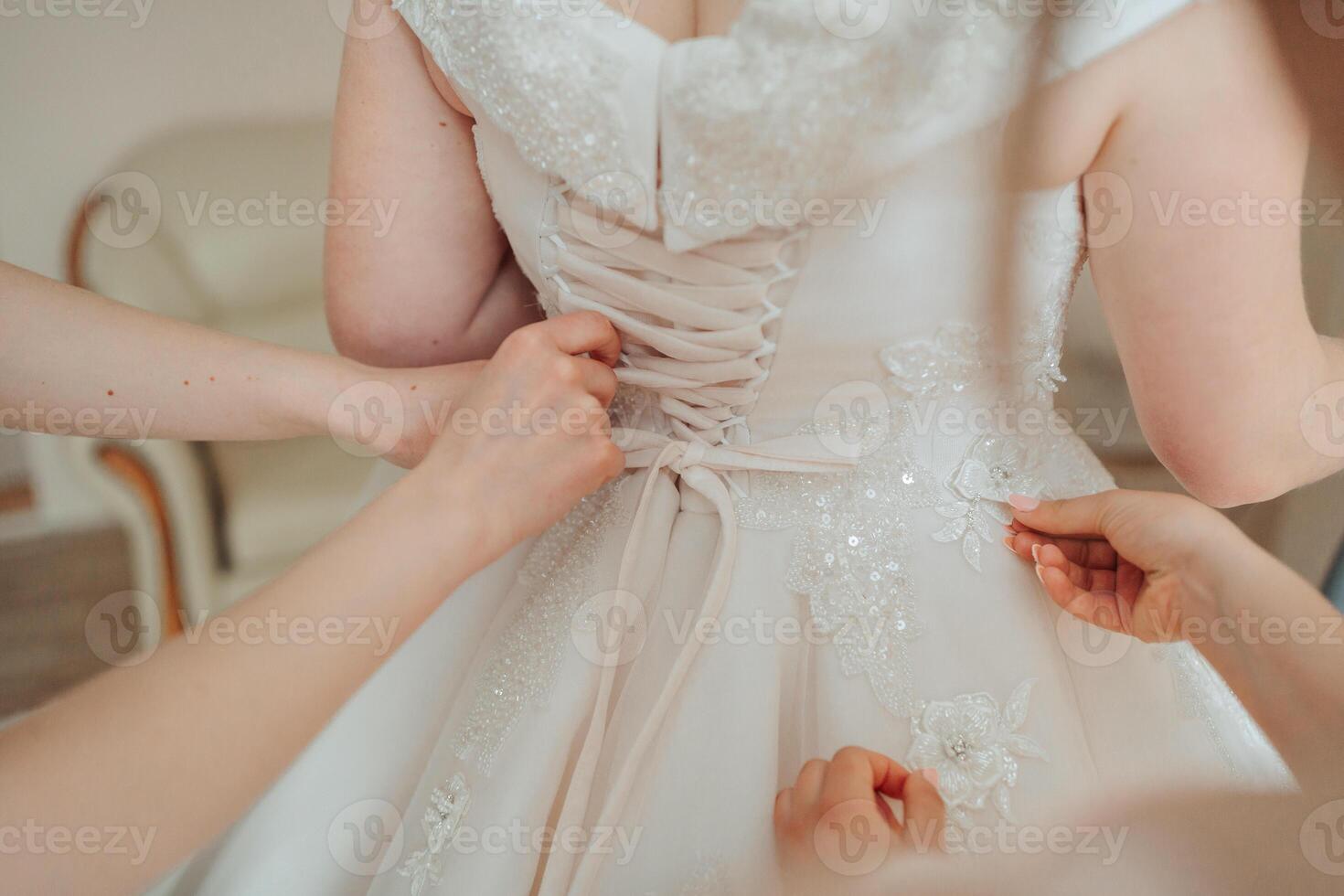 Three women are getting ready for a wedding. One of them is adjusting the lace on a white dress photo