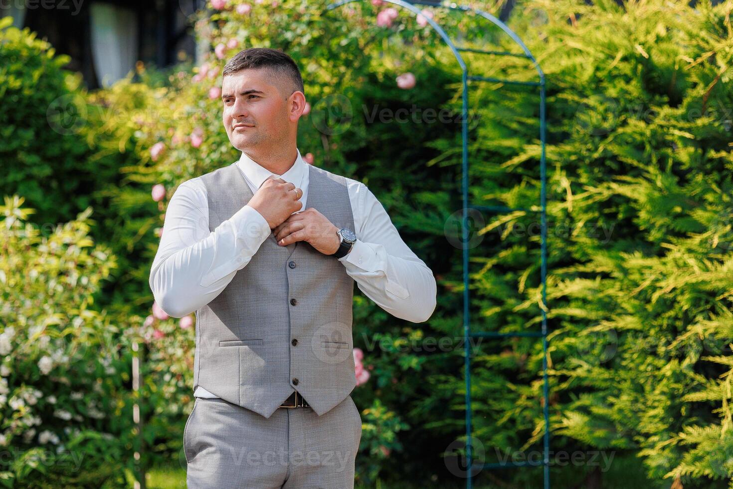 A man in a suit and vest stands in front of a green archway. He is adjusting his tie and he is in a professional setting photo