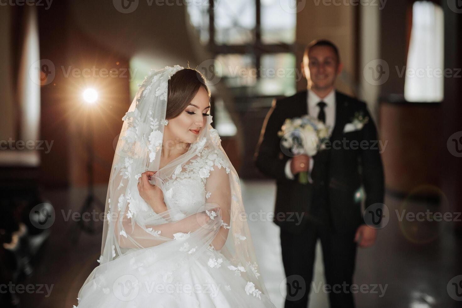 A bride and groom are standing in front of a camera. The bride is wearing a veil and a white dress. The man is holding a bouquet of flowers photo