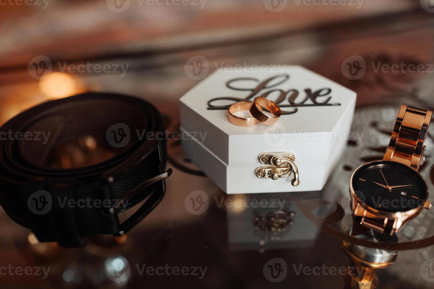 A white box with the word love written on it sits on a table. Inside the box are two gold wedding rings. A black belt is also on the table photo