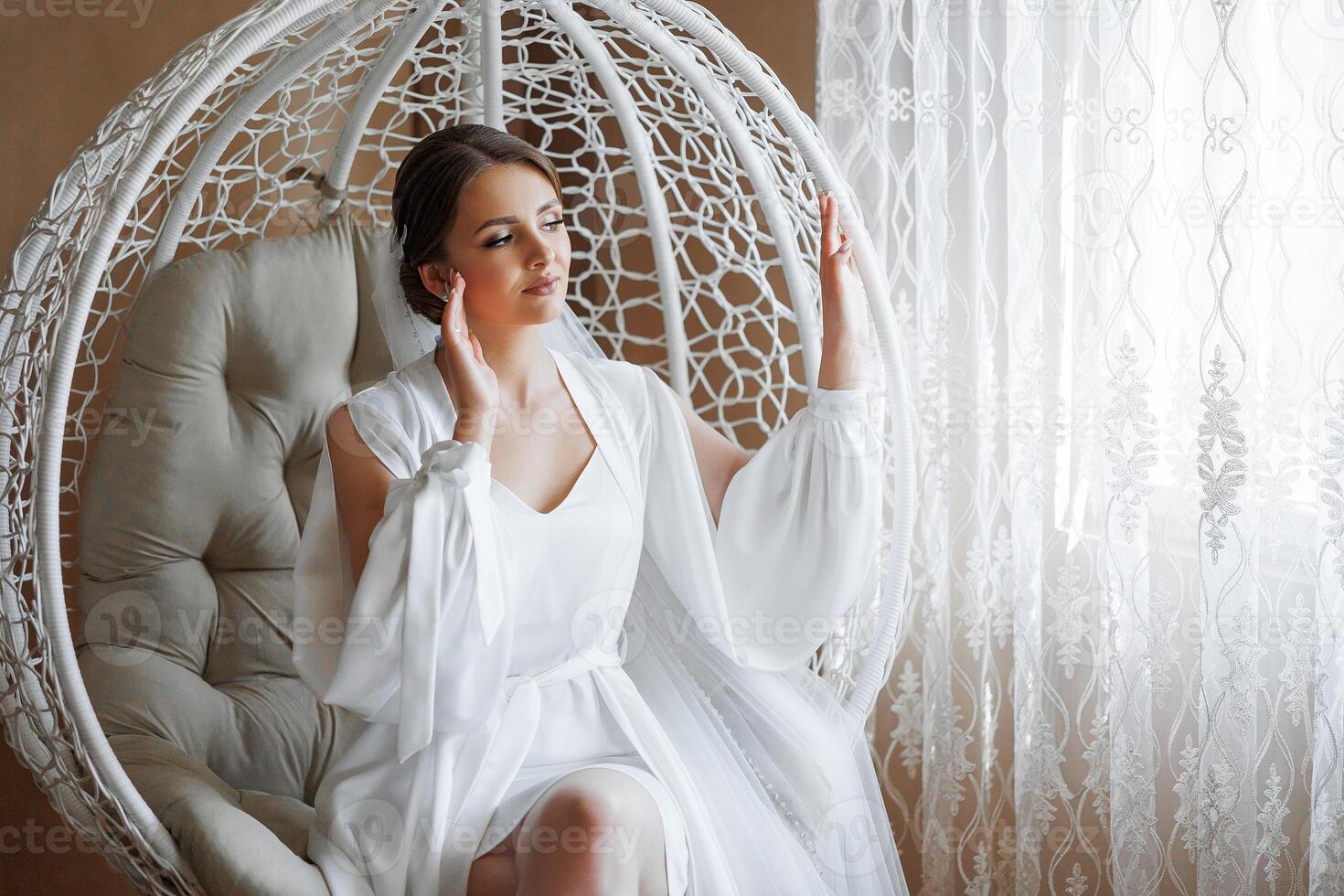 A woman in a white gown sits in a white chair. She is looking out the window photo