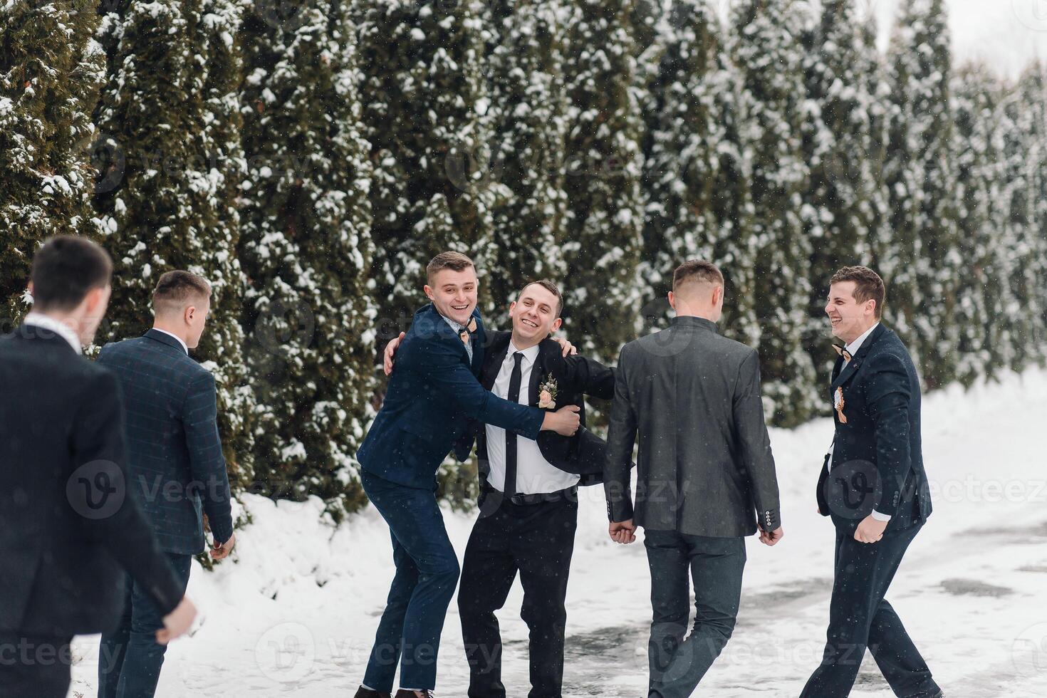 A group of men are standing in the snow, dressed in suits and ties. They are laughing and hugging each other, enjoying the winter weather photo