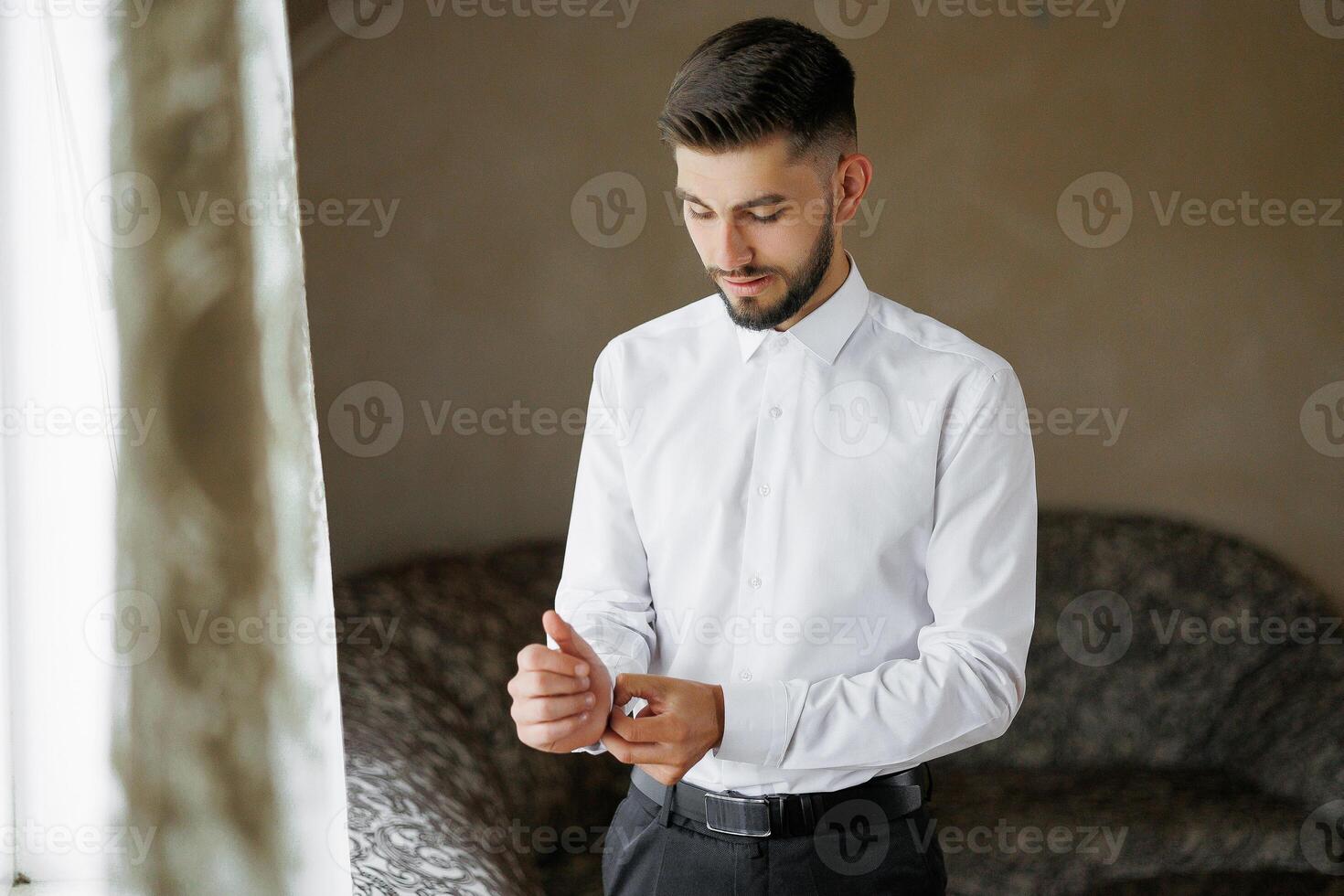 A man in a white shirt is getting ready to dress up. He is standing in front of a couch and a window photo