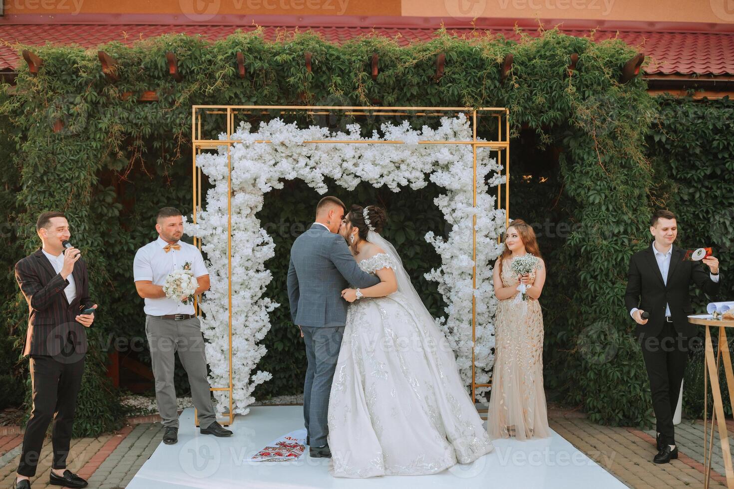 A bride and groom are kissing under a white arch. The bride is wearing a long white dress and the groom is wearing a suit. There are several other people in the background photo