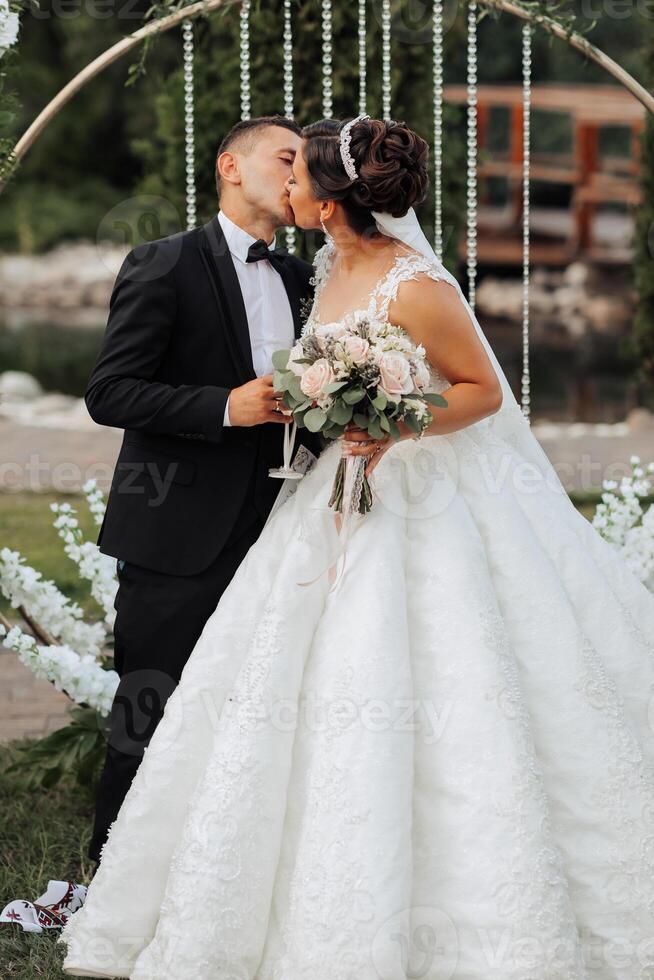 A bride and groom are kissing in front of a wedding arch. The bride is holding a bouquet of flowers photo