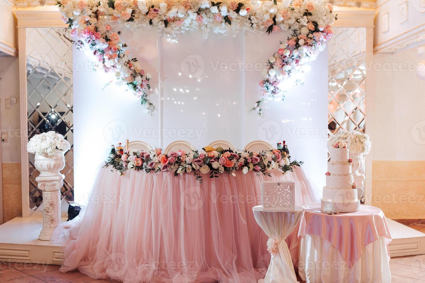 A wedding reception with a pink tablecloth and floral decorations. A cake is on the table and there are chairs around it photo