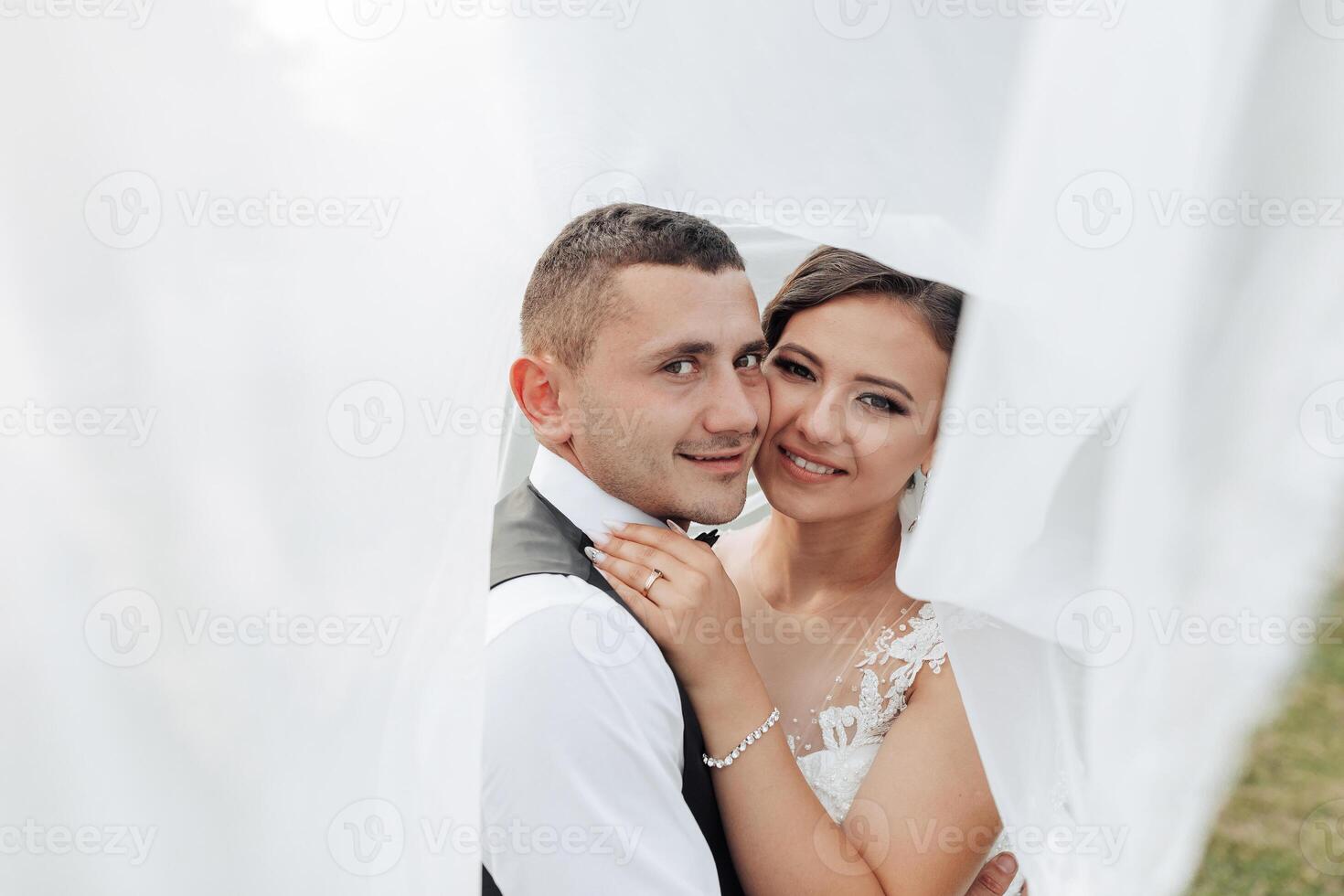 A bride and groom are posing for a picture under a white veil. The bride is wearing a white dress and the groom is wearing a black suit. They are both smiling and looking at the camera photo