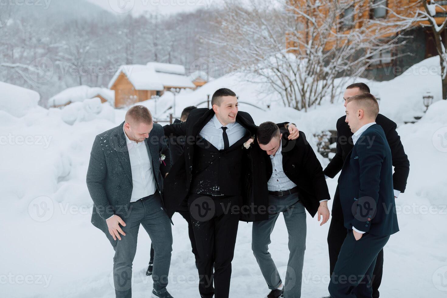 A group of men in suits are posing for a picture in the snow. They are all smiling and seem to be enjoying themselves. Scene is lighthearted and fun, as the men are embracing each other photo