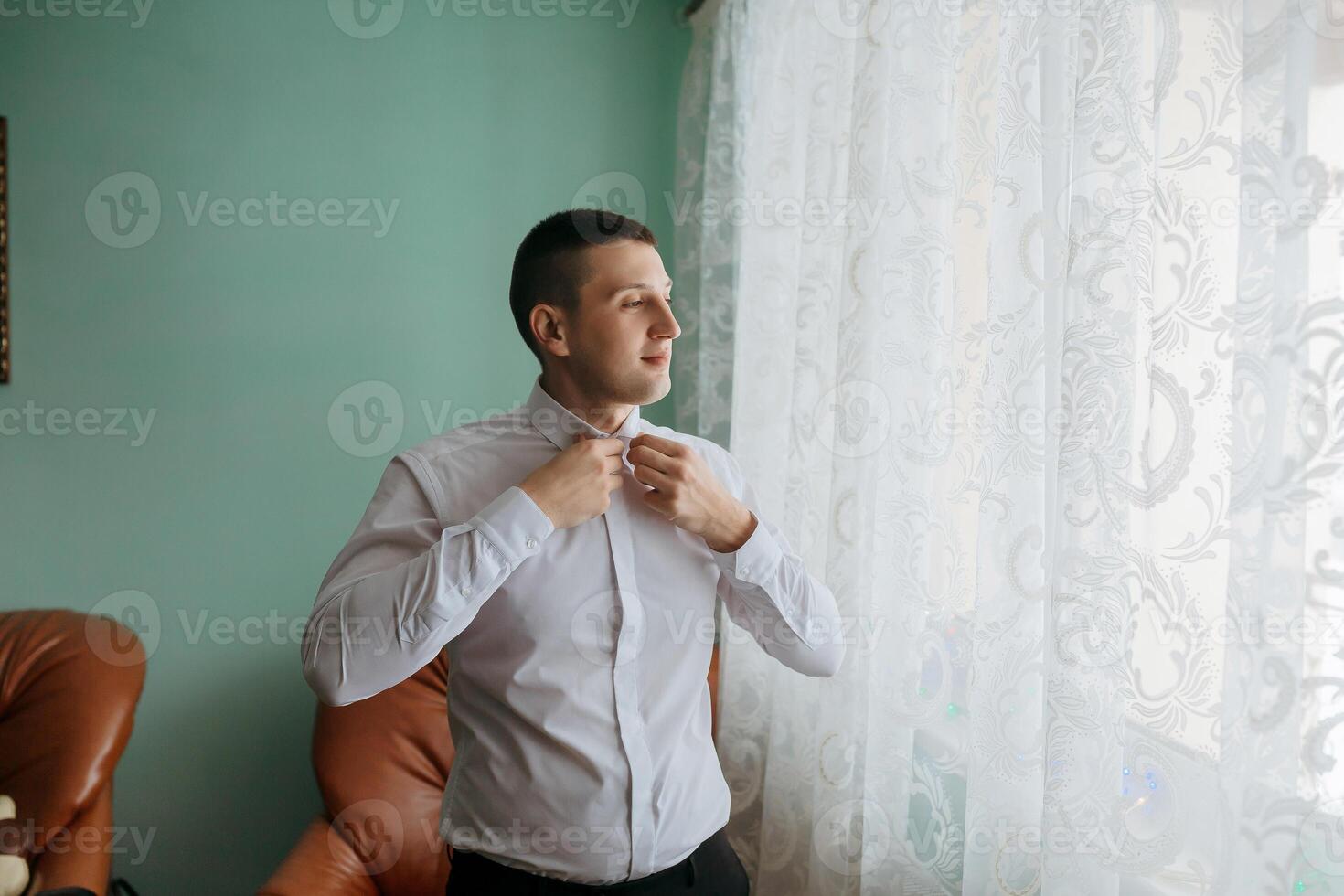 A man in a white shirt is getting dressed in front of a window. The room is decorated with a green wall and a brown chair photo