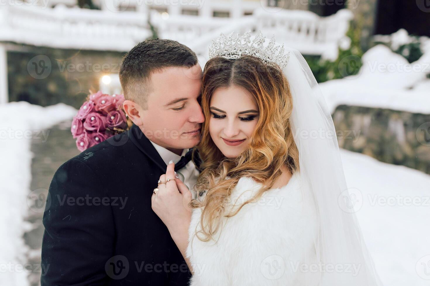 A bride and groom are posing for a picture in the snow. The bride is wearing a white fur stole and a tiara photo