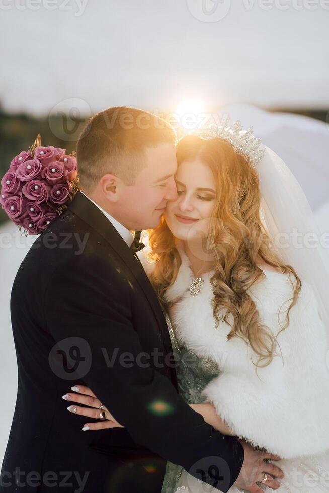 A bride and groom are embracing each other in the snow, with the bride wearing a white fur coat and the groom holding a bouquet of pink flowers photo