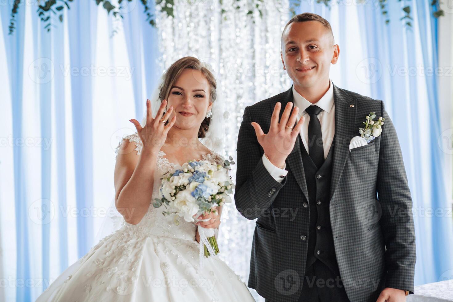 A bride and groom are standing in front of a blue curtain, with the bride holding a bouquet photo