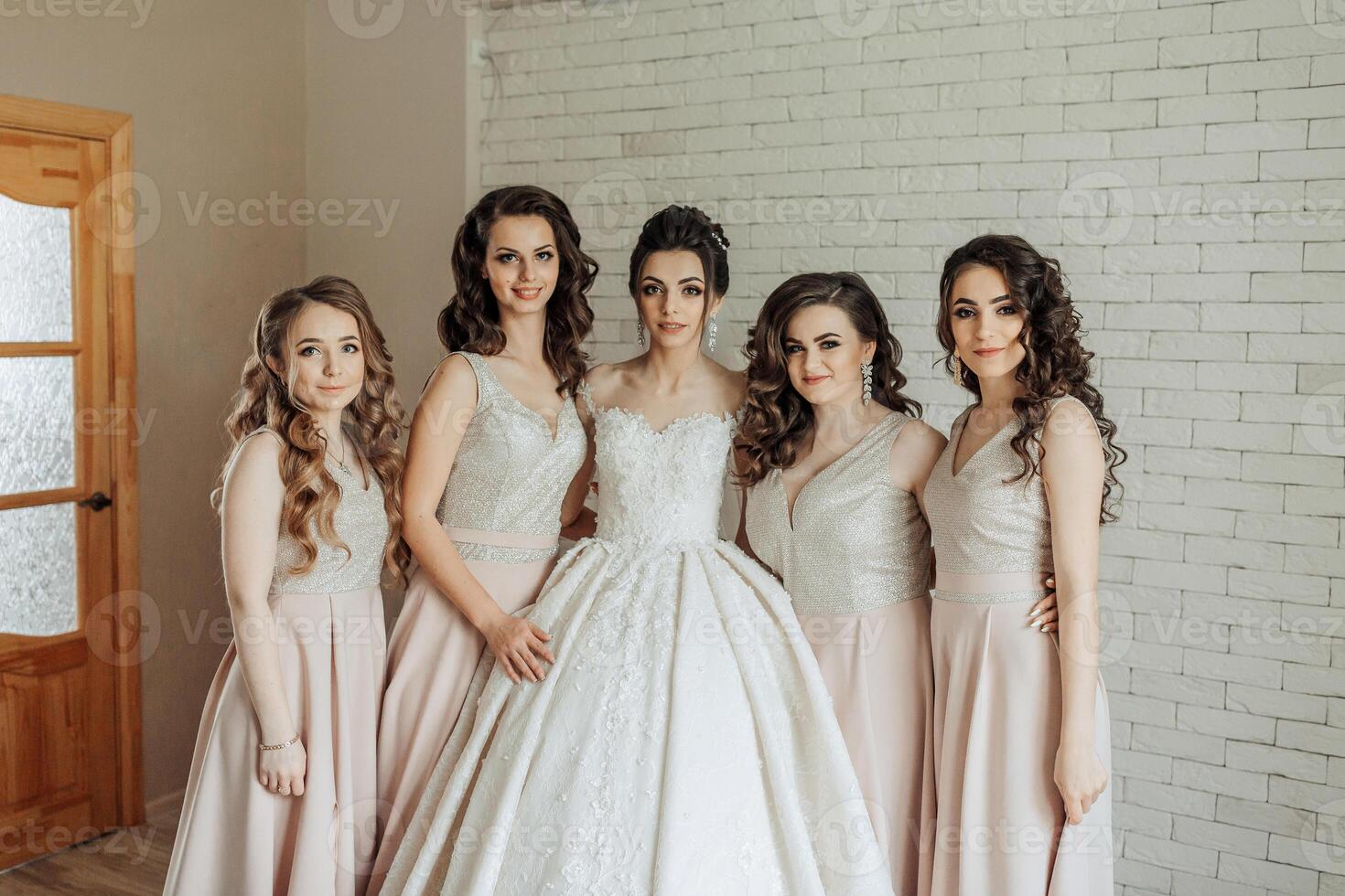 A group of five women are posing for a picture in front of a white wall. They are all wearing dresses and are smiling for the camera. Scene is happy and celebratory photo