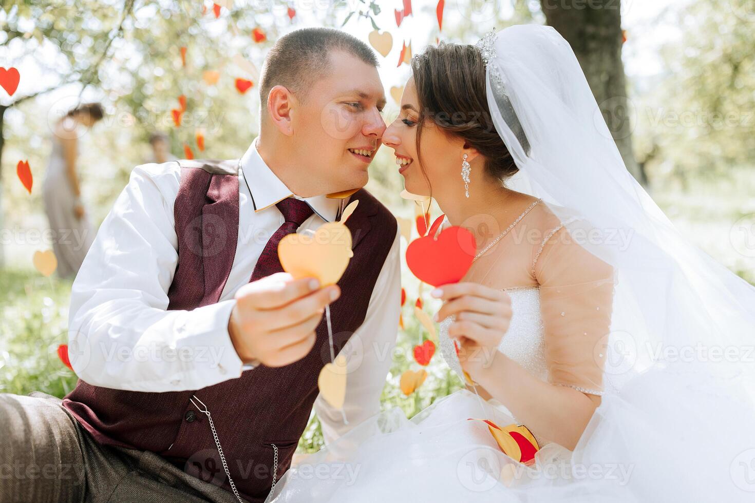A bride and groom are sitting in a field with red hearts falling around them. The bride is holding a red heart and the groom is holding a red heart as well photo
