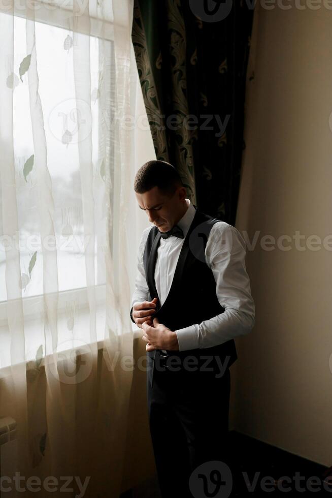 A man in a suit is standing in front of a window, adjusting his tie. Concept of formality and attention to detail, as the man takes the time to ensure his appearance is polished and professional photo