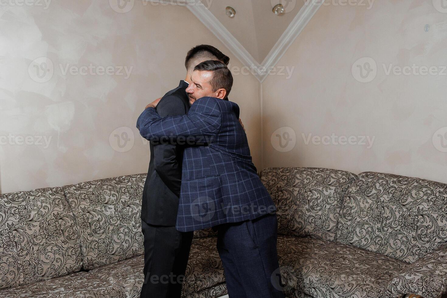 Two men hug each other in a living room. One of them is wearing a suit. Scene is warm and friendly photo