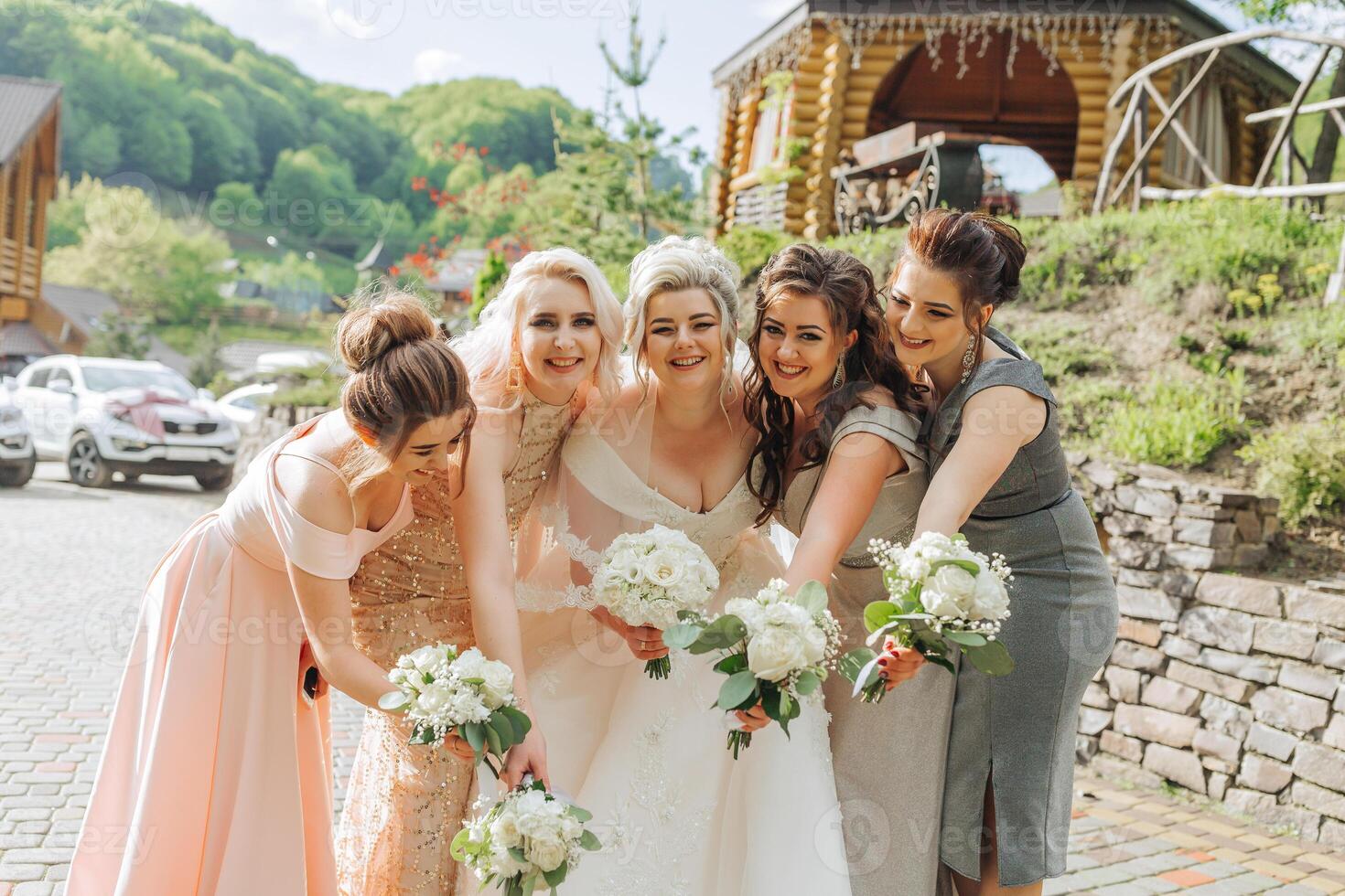 A group of women are posing for a photo, with one of them wearing a wedding dress. They are all holding white flowers, and there are several cars in the background. Scene is joyful and celebratory photo
