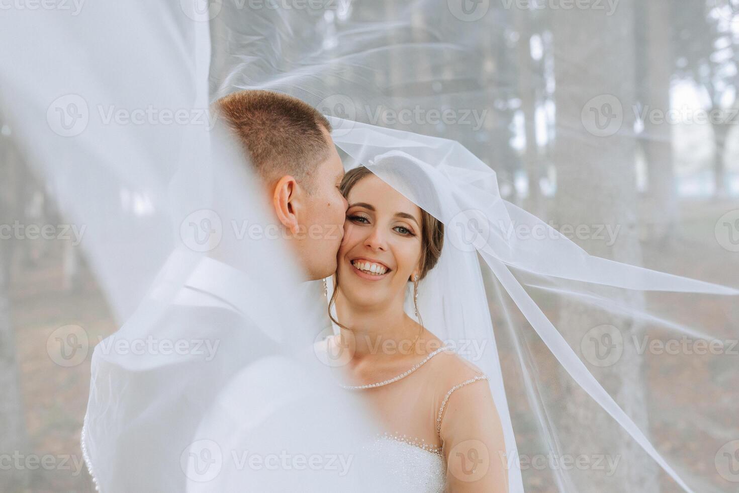 A bride and groom are kissing under a veil. The bride is wearing a white dress and the groom is wearing a white shirt photo