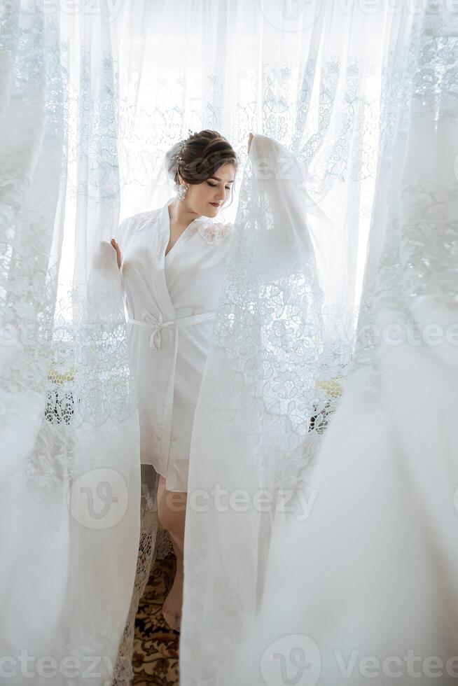 A woman in a white robe stands in front of a curtain, with a lace veil on her head photo