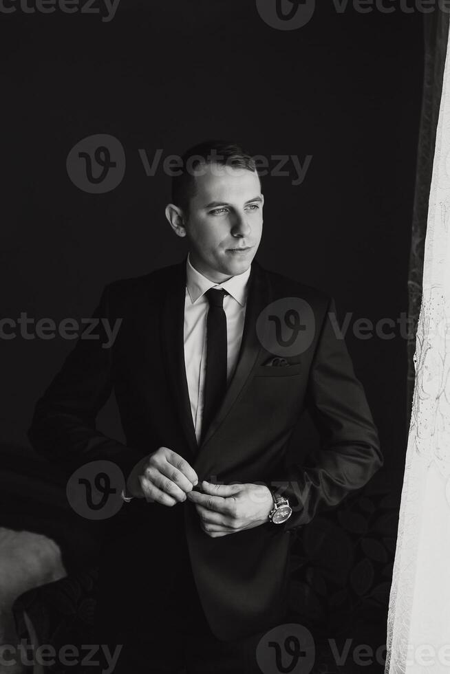 A man in a suit is standing in front of a window, adjusting his tie. Concept of formality and professionalism, as the man is dressed in a suit and tie photo