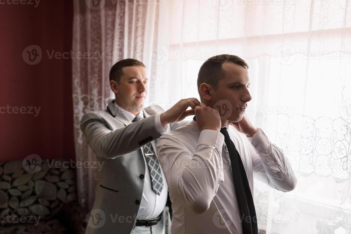 Two men are getting dressed up, one of them is wearing a tie. The man in the suit is helping the other man tie his tie photo